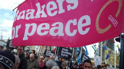 In a busy crowd Quakers hold up a hot pink 'Quakers for peace' banner alongside 'Stop Bombing Syria' placards with a scaffolded Houses of Parliament in the background