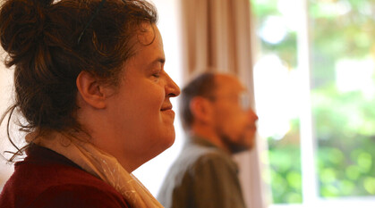 Close up of a Quaker with eyes closed in meeting for worship. The colours are soft and warm with ight coming through the window with green foliage outside.