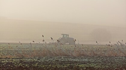 A tractor in a misty field, surrounded by tall grasses, with hills in the distance.