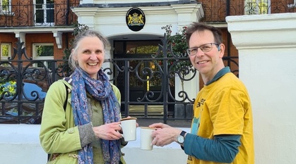 Two people standing outside an embassy holding cups of coffee and stroopwafels