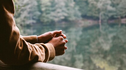 A pair of hands lean on a rail at the side of a body of water, which reflects the trees growing by its side.
