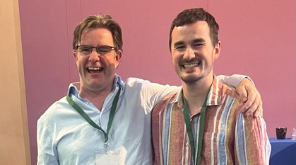 Two men stand together, laughing; one has his arm around the other's shoulders.