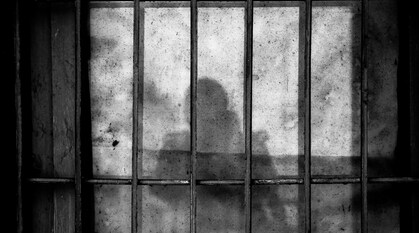 A black and white photo of a dirty window behind bars with the shadow of a person in view